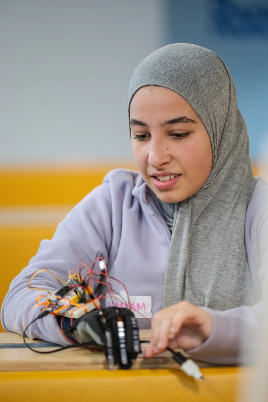 Maryam working at a bench on the Sign Scribe glove's circuit board, holding a tool, with the wired glove on the table in front of her