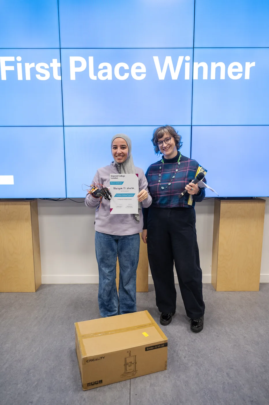 Two students standing in front of a large screen reading First Place Winner, one holding a certificate and a wired glove, with a boxed 3D printer on the floor in front of them